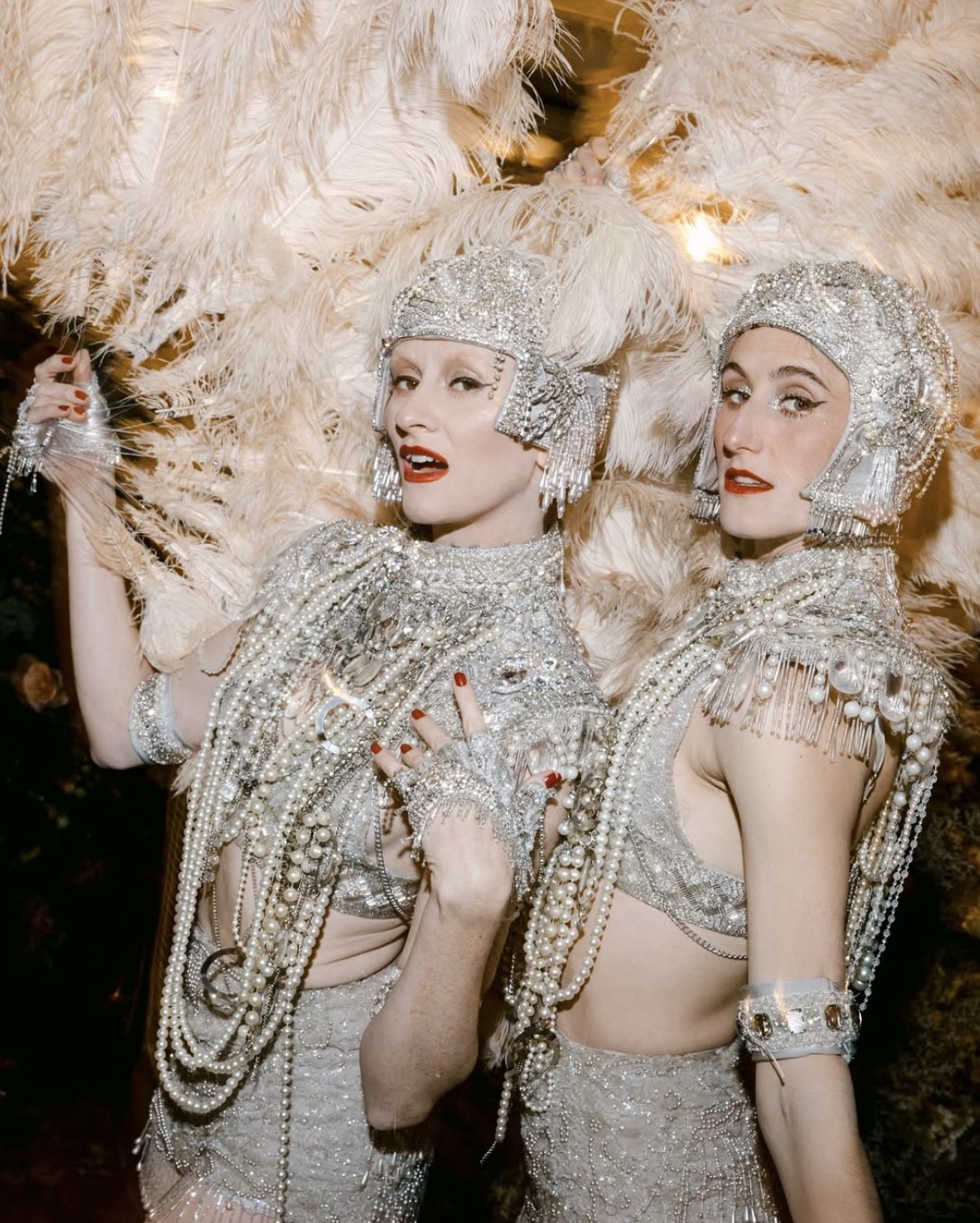 Two performers in silver crystal-beaded corsets and fitted helmet headpieces trimmed with crystal fringe, holding large cream ostrich feather fans