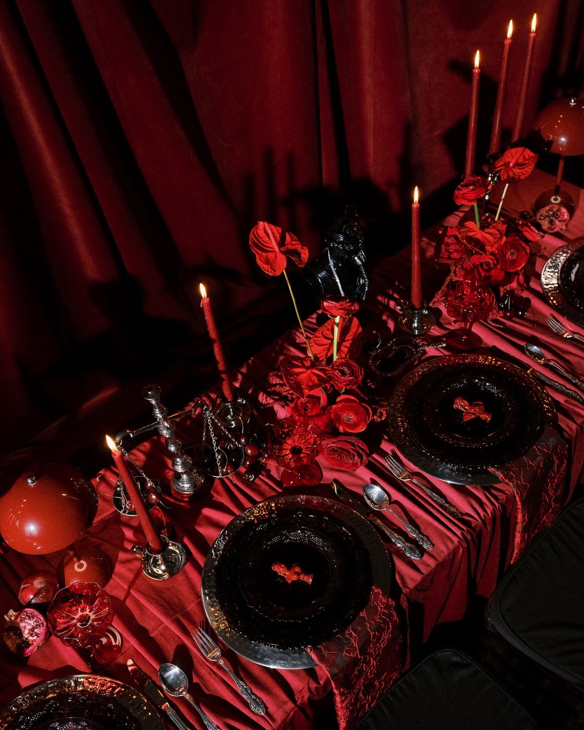 A banquet table dressed in deep red fabric, burning taper candles, red flowers, cut pomegranates and a black bust against crimson drapery
