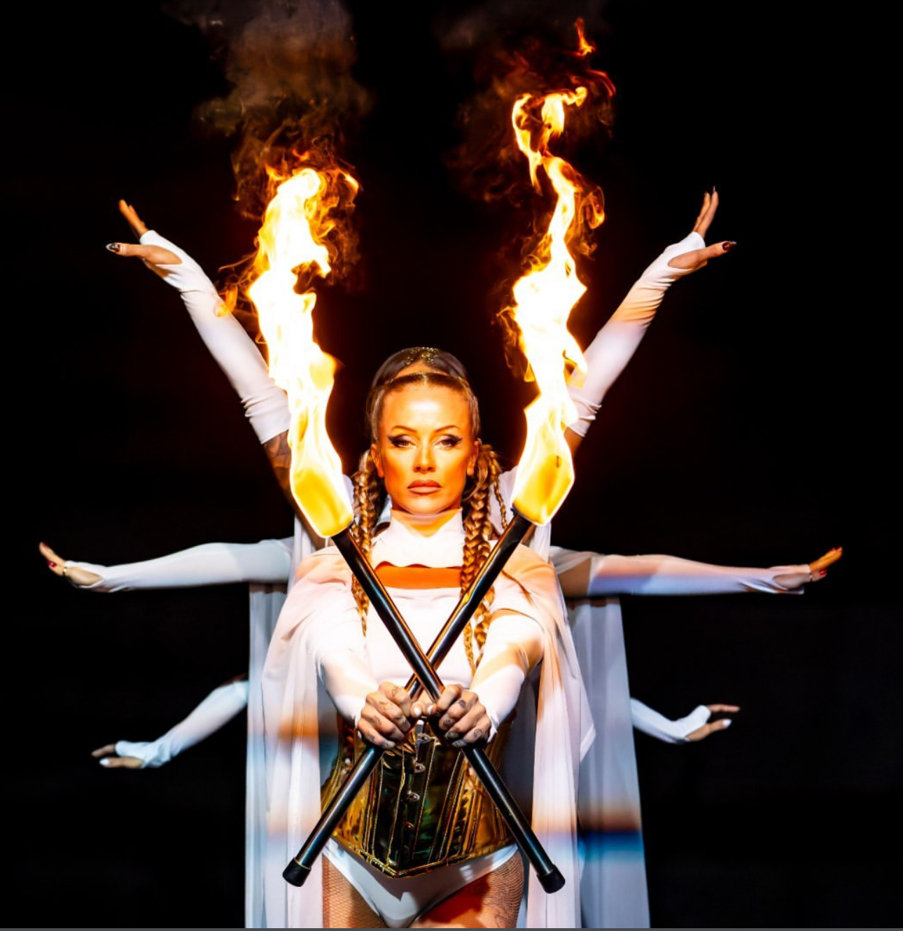 Fire performer holding crossed flaming torches with dancers behind her