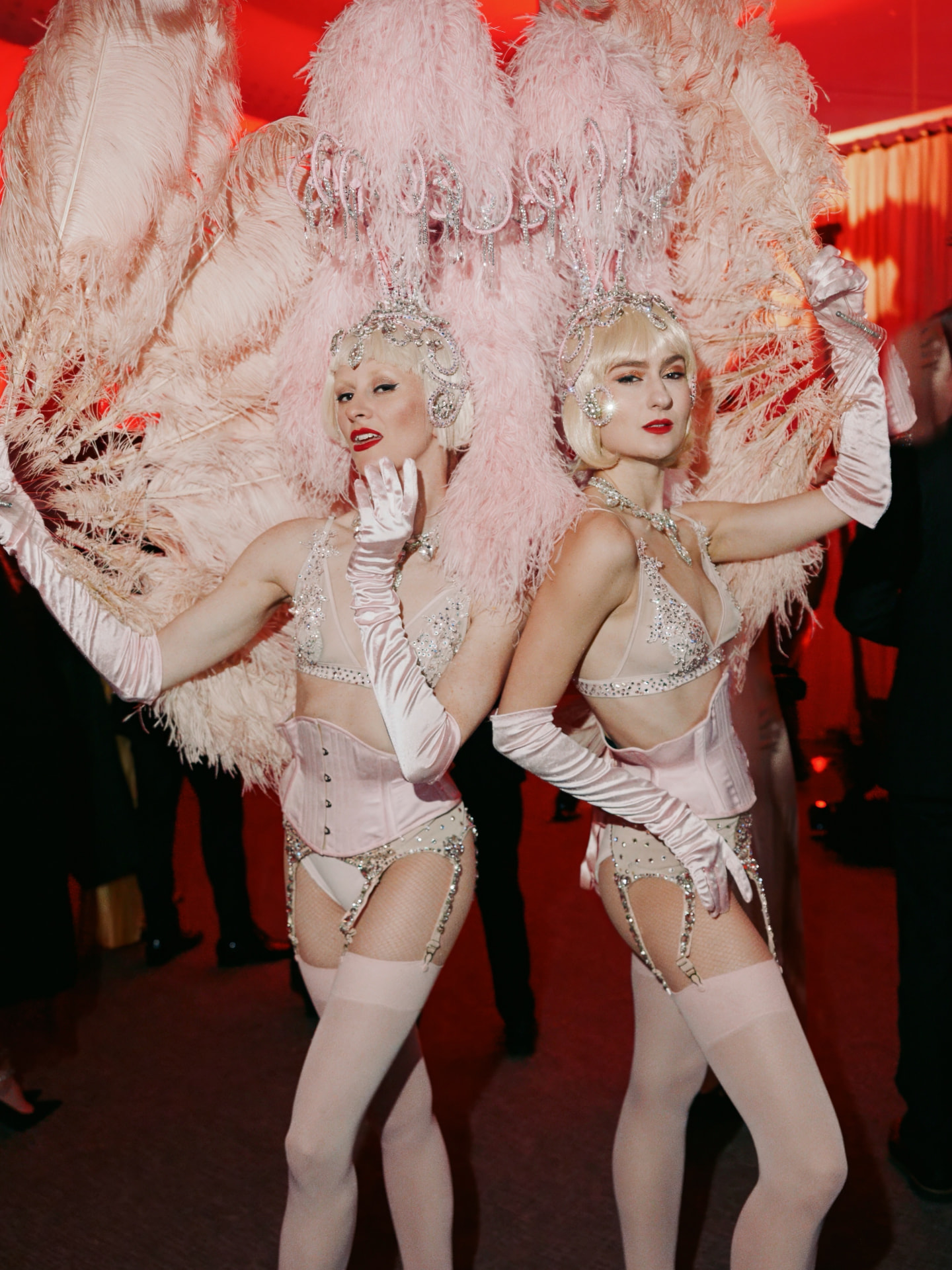 Two showgirls in blush pink feather headdresses and crystal corsets at a red-lit gala