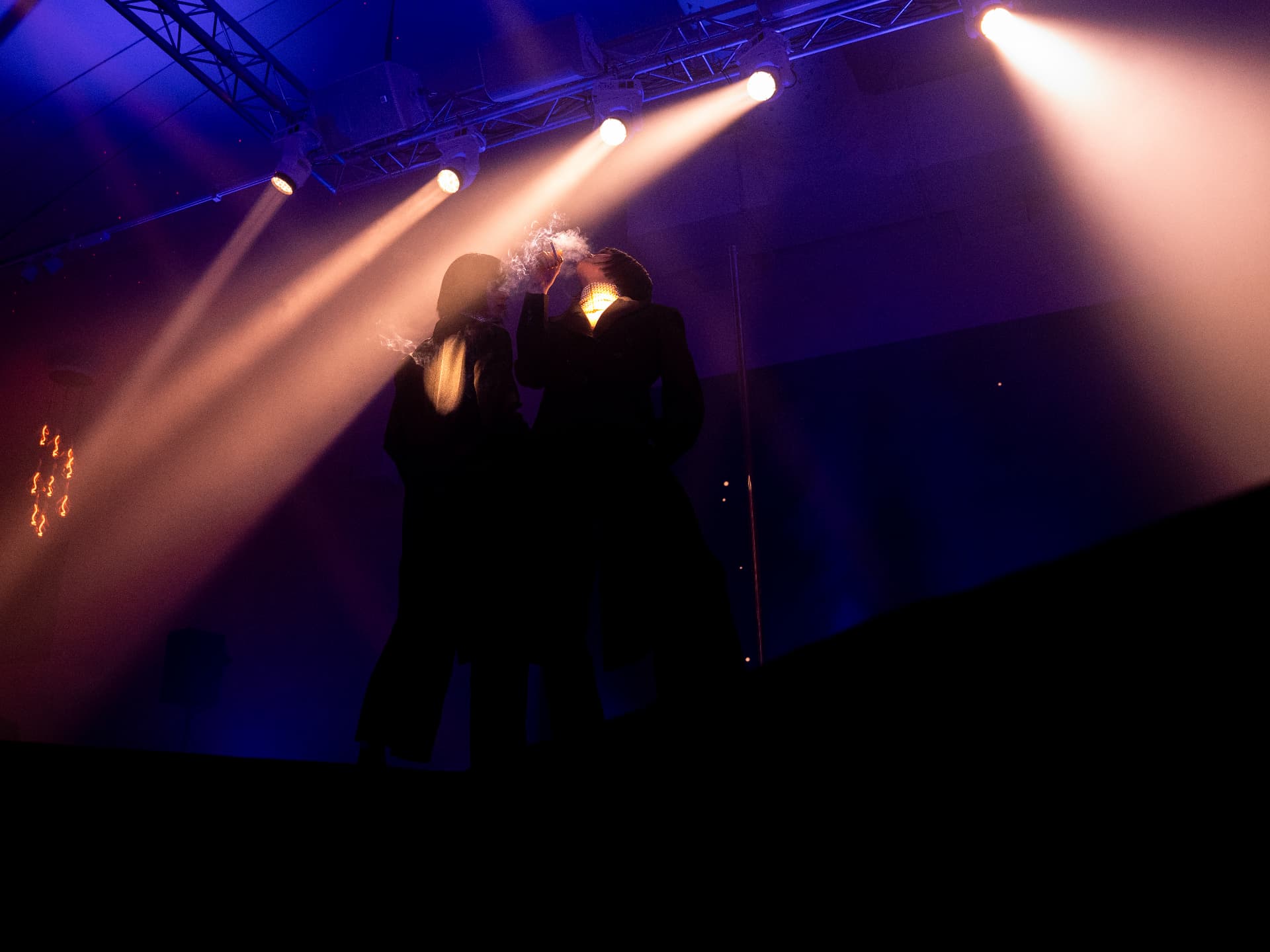 Two performers in dark tailored stage coats on a raised platform, one exhaling illuminated smoke beneath warm spotlight beams from an overhead truss