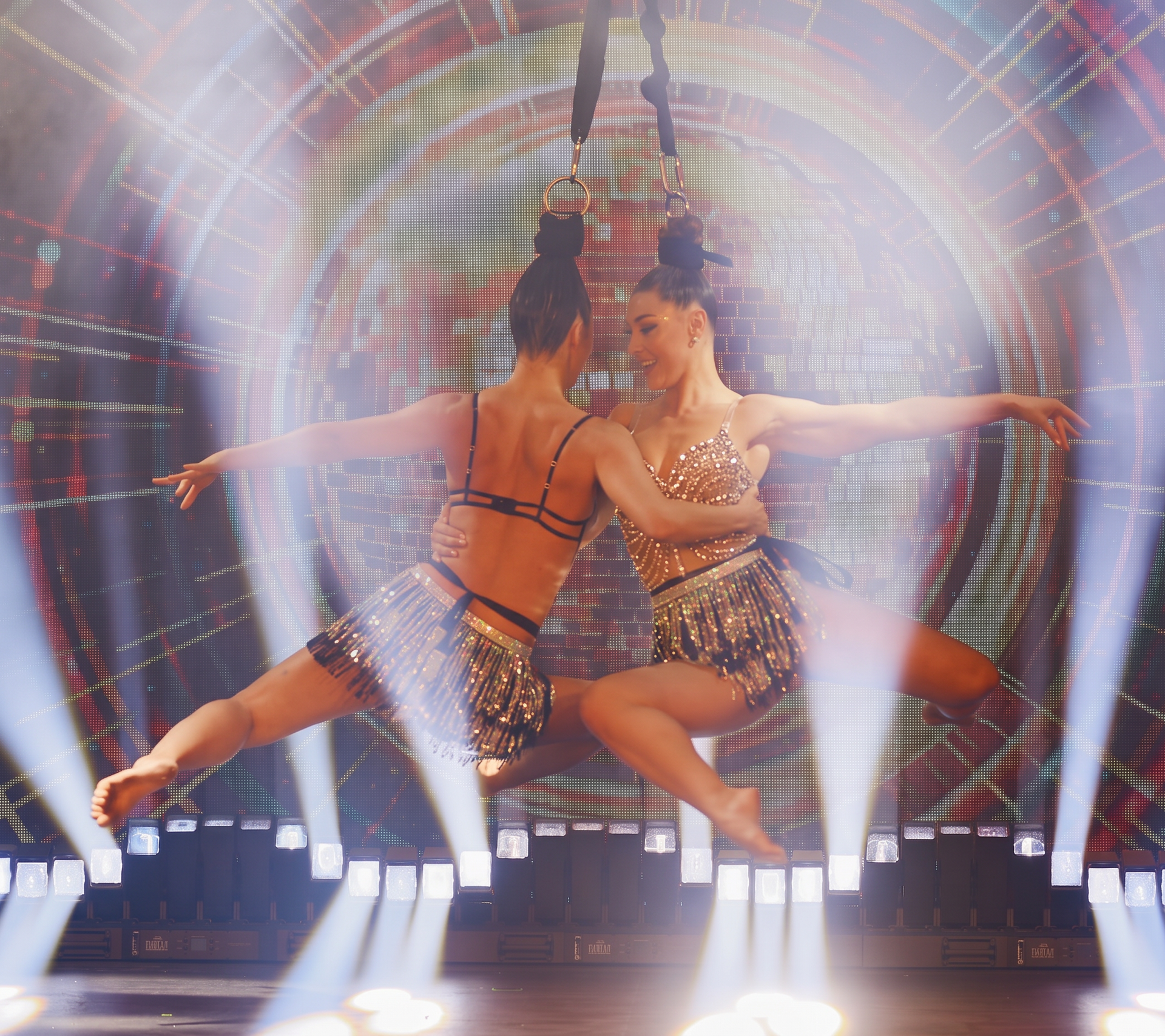Two aerial performers suspended on straps, facing one another mid-air before a circular LED backdrop
